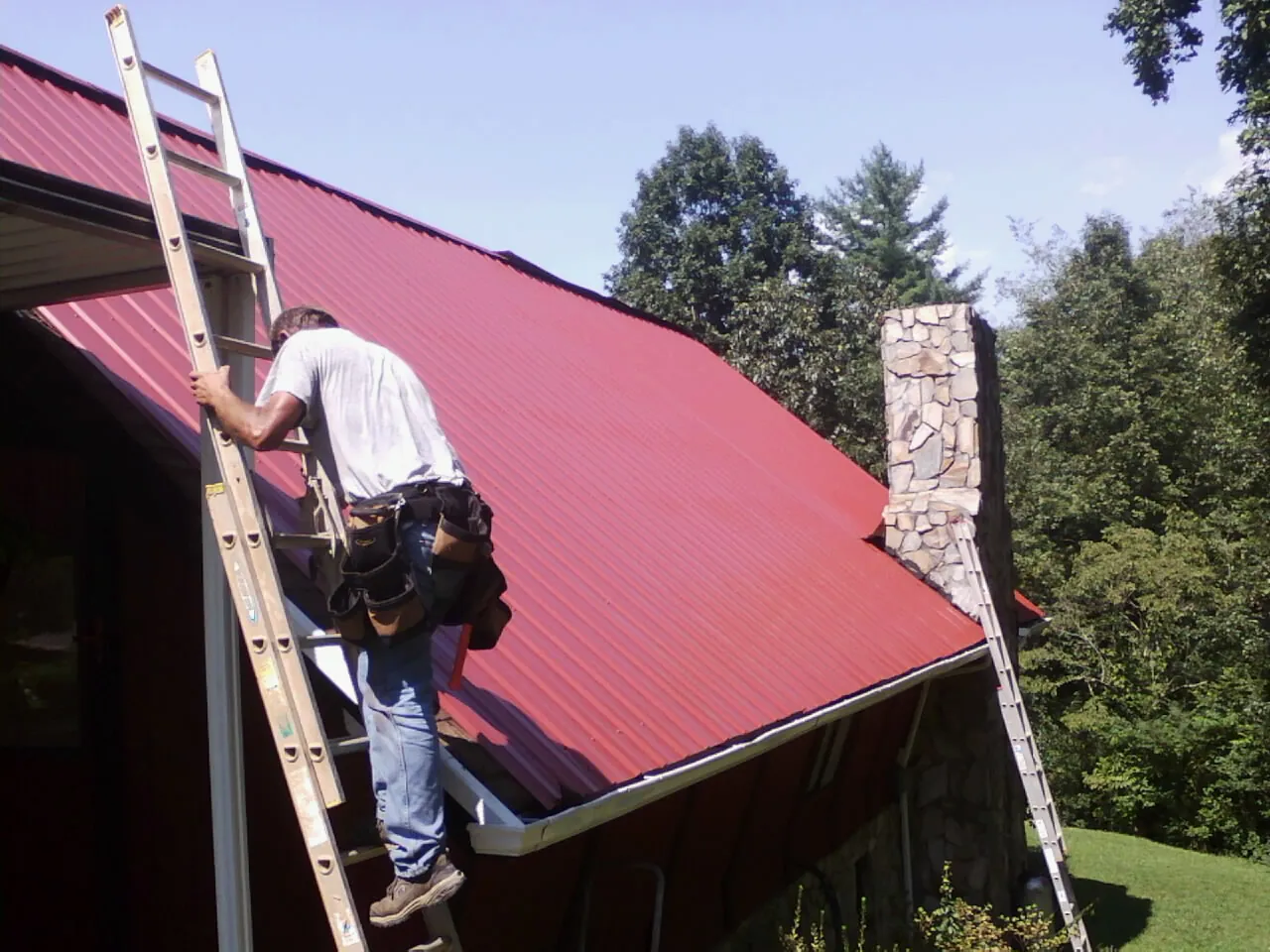 Roofer on a ladder beside a red metal roof and stone chimney — Soffit & Fascia Repair by Roofing Brothers in Fromberg